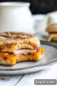 Easy Make Ahead Breakfast Sandwiches: Freezer-Friendly Eggs, Veggies, Canadian Bacon, and Cheese on English Muffins. Perfect for quick breakfasts, lunches, or meal prep for kids and adults! A delicious make-ahead breakfast sandwich with egg, cheese, and Canadian bacon, with a bite taken out, resting on a gray plate on a white kitchen counter.