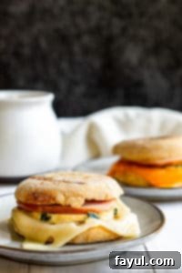 Make Ahead Breakfast Sandwiches: Fresh, delicious, and ideal for meal prep. Featuring eggs, Canadian bacon, cheese, and optional veggies on English muffins. A straight-on shot of a make-ahead breakfast sandwich on a gray plate, with a second sandwich blurred in the background, highlighting the fresh ingredients.
