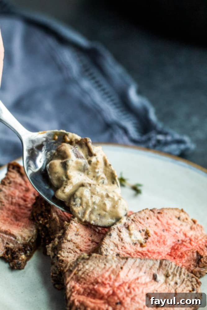 Silver spoon pouring mushroom sauce over sliced peppercorn steak on a gray plate.