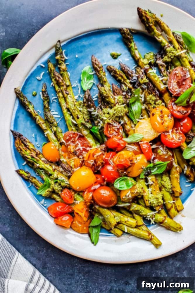 This Balsamic Oven Roasted Asparagus recipe is the perfect side dish! This easy vegetarian recipe is topped with fresh tomatoes, basil, Parmesan cheese, and homemade basil pesto. This side pairs great with chicken, steak, or soup and salad for a healthy pairing. #recipe #vegetarian #healthy Overhead shot of finished balsamic roasted asparagus recipe on a platter on a blue counter, garnished with fresh basil and tomatoes.