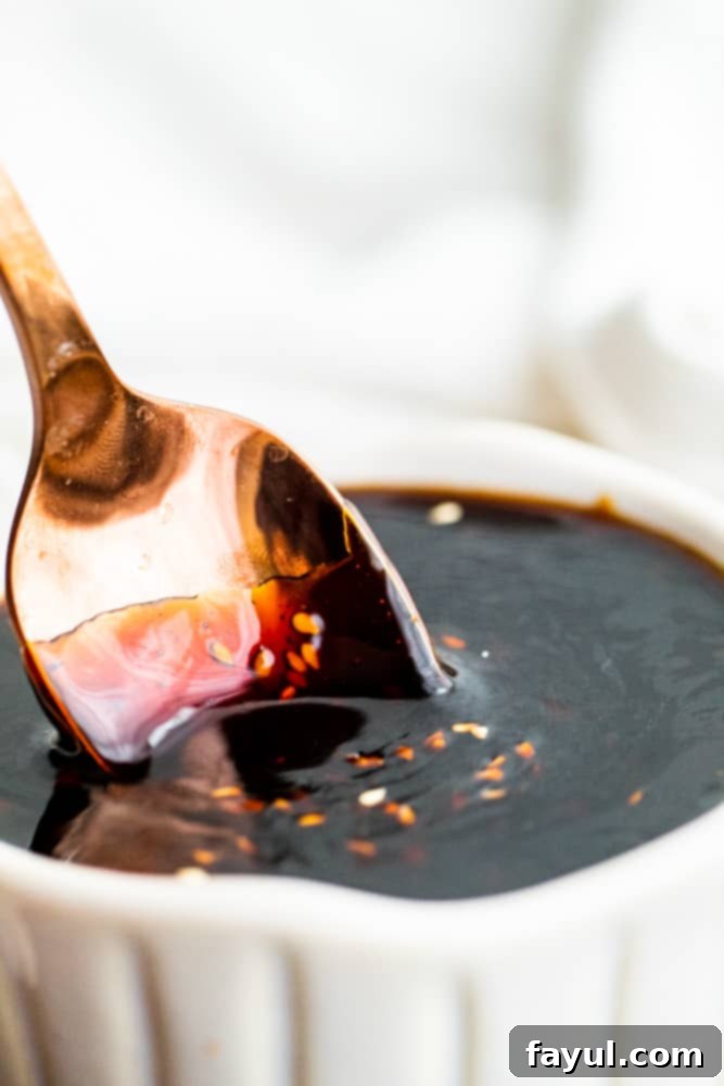 This Easy Homemade Teriyaki Sauce recipe is a great DIY version of this Asian favorite. This sauce pairs perfect with stir fry, chicken, beef and vegetables. This simple Japanese sauce is ready in about 20 minutes and has the classic teriyaki sauce flavor that we all know and love. #TeriyakiSauce #recipe #Asian A golden spoon scooping a portion of rich, glossy homemade teriyaki sauce from a white serving dish, highlighting its thick and sticky texture.