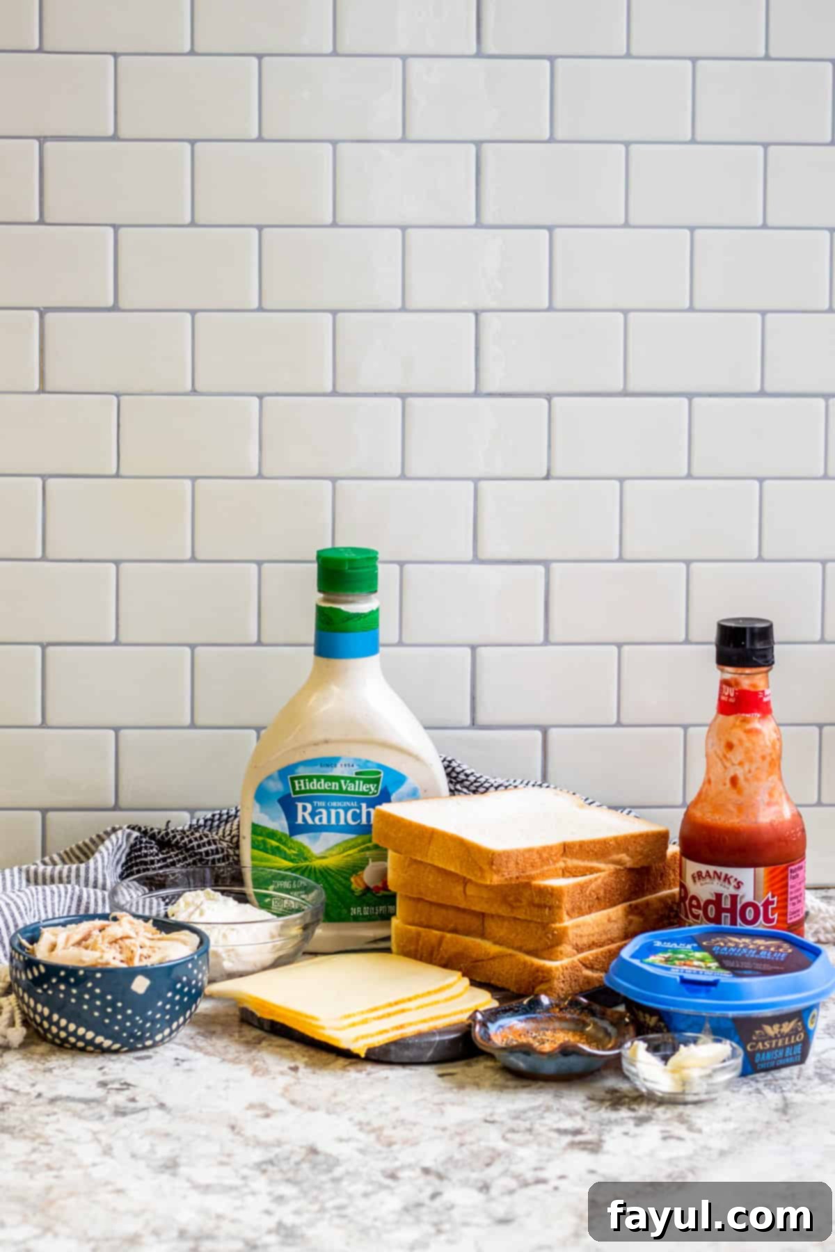 An inviting overhead shot of various fresh ingredients for buffalo chicken grilled cheese arranged in small bowls and containers on a rustic kitchen counter, ready for preparation.