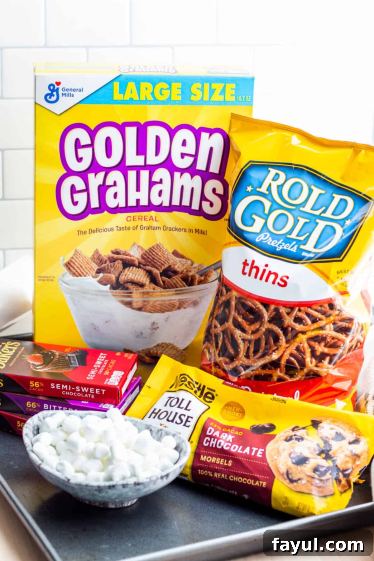 A flat lay photograph showcasing all the ingredients for a delicious no-bake S'mores Snack Mix laid out on a white surface: golden grahams cereal, mini marshmallows, pretzels, dark chocolate chips, and melting baking chocolate.