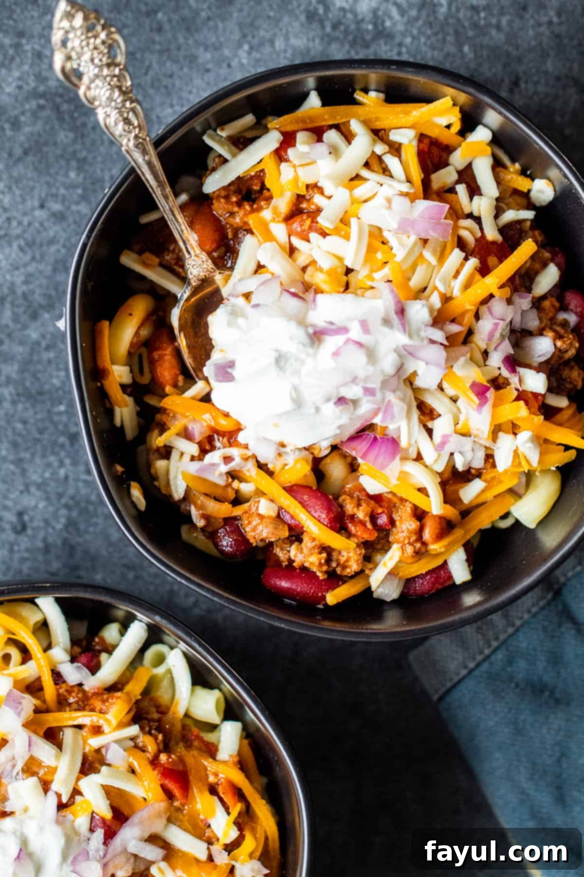 Overhead shot of two black bowls with chili mac and cheese inside on dark blue counter, garnished with cheese and sour cream.