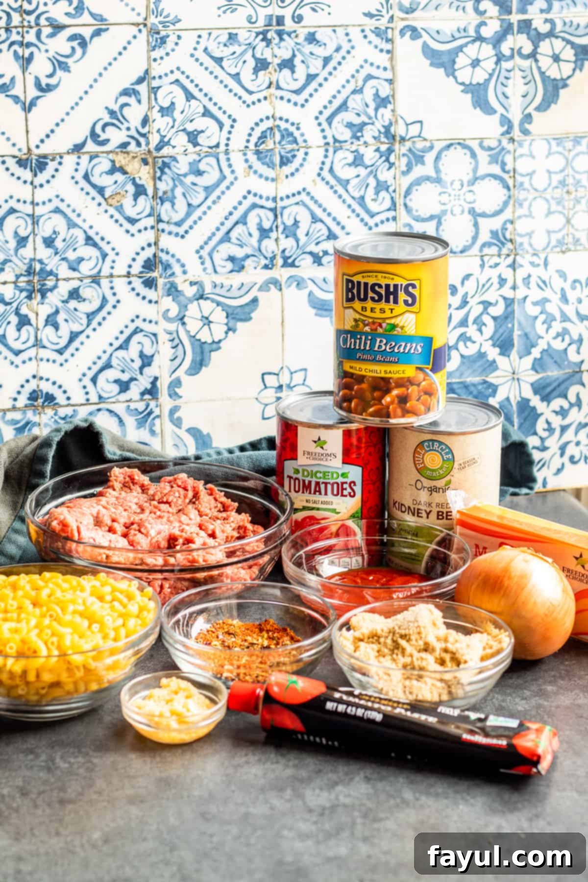 Chili ingredients laid out on table in bowls, containers and plates, including meats, spices, and canned goods.