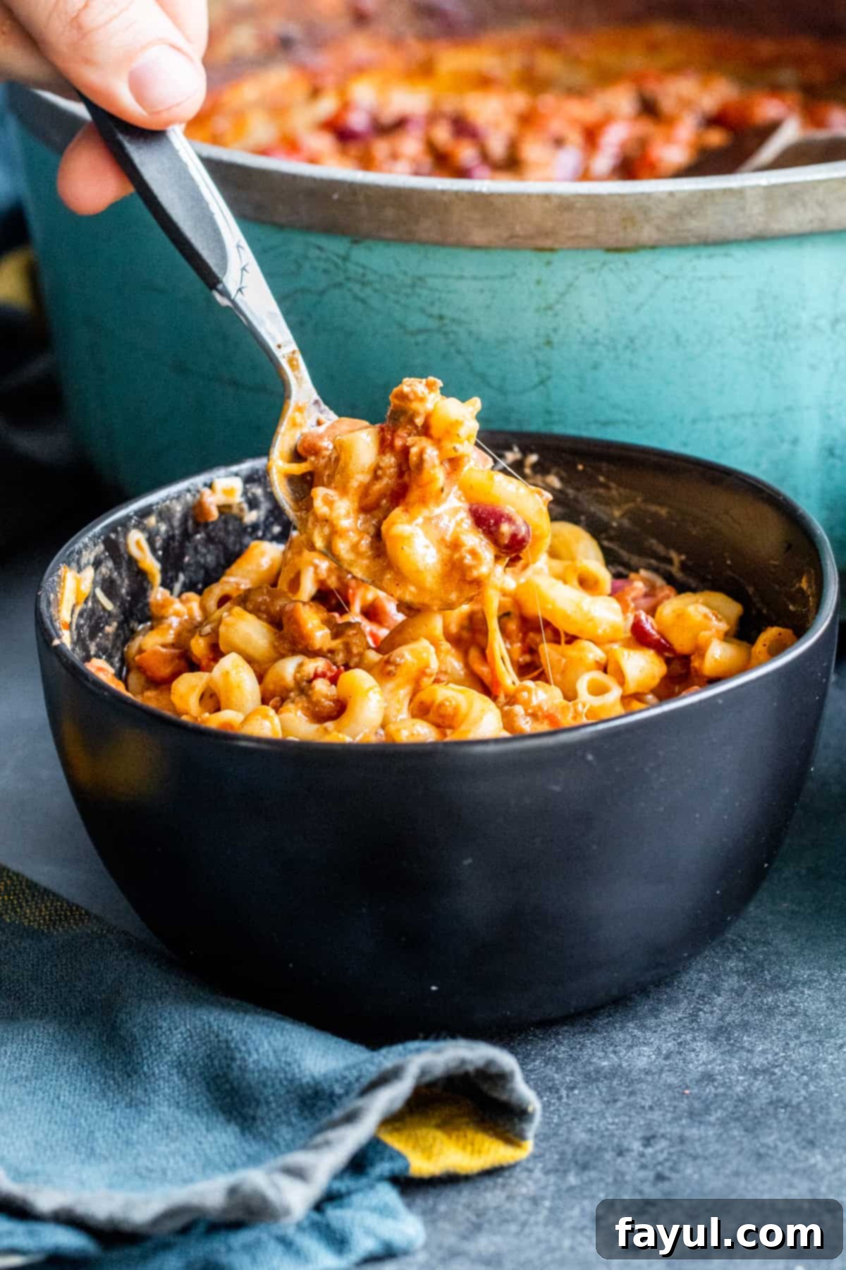 Spoon scooping up chili with noodles from black bowl, showing melted cheese.