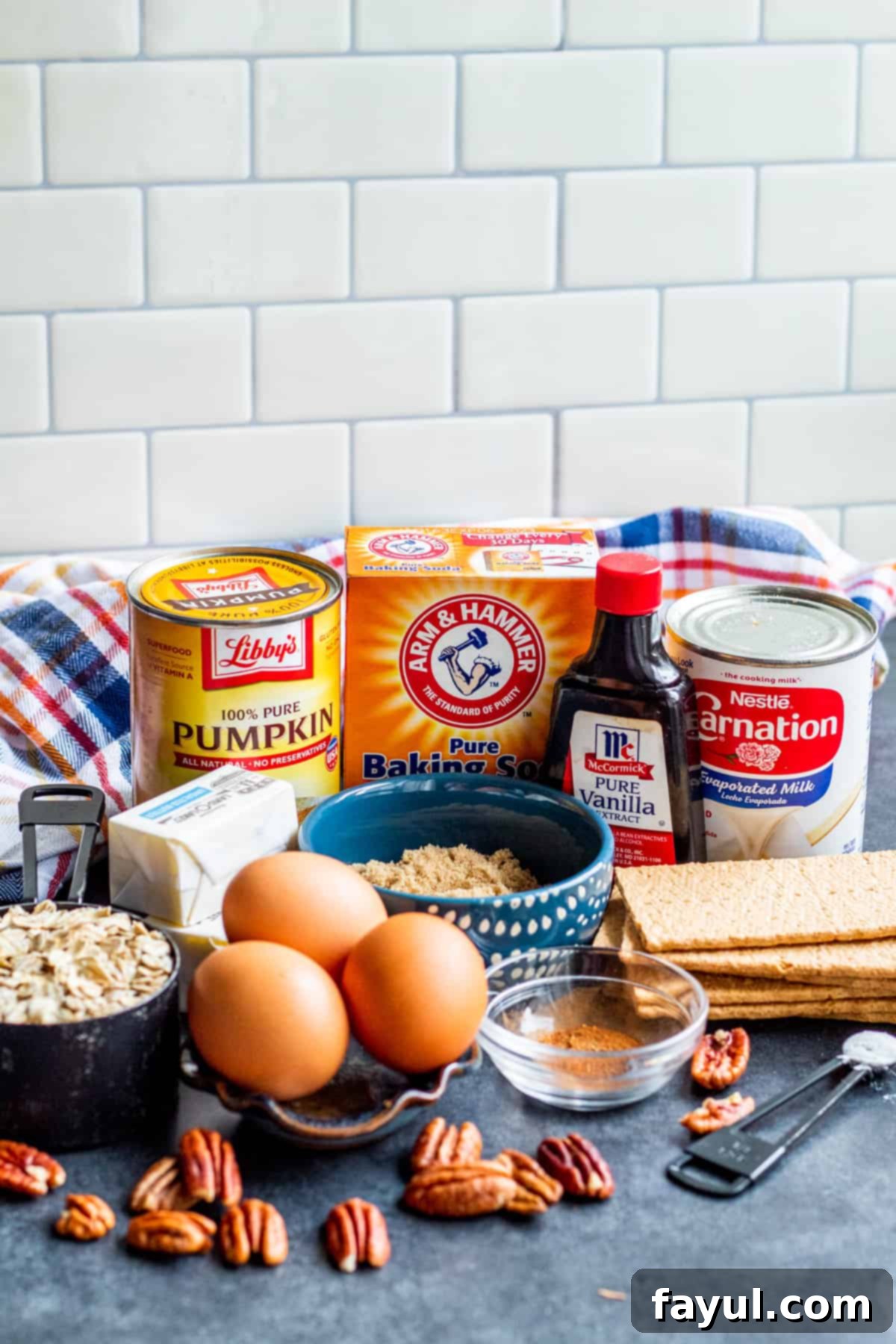 Irresistible Pumpkin Crisp featuring Pecan Oat Streusel 3 An organized display of pumpkin crisp recipe ingredients. Small bowls contain canned pumpkin, brown sugar, eggs, evaporated milk, vanilla extract, pumpkin pie spice, and sea salt for the filling. Separately, graham crackers, old-fashioned oats, more brown sugar, baking powder, baking soda, and pecans are arranged for the topping, alongside a stick of butter. All items are neatly arranged on a rustic blue wooden table, ready for preparation.