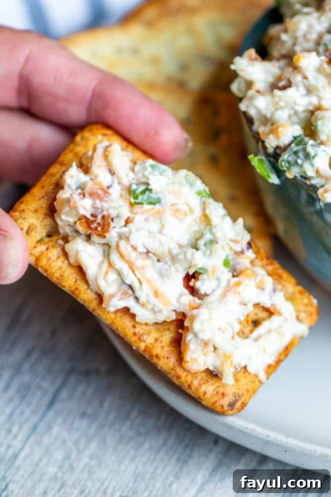A hand gracefully picking up a cracker generously topped with the creamy Neiman Marcus Million Dollar Dip, held over a clean white plate.