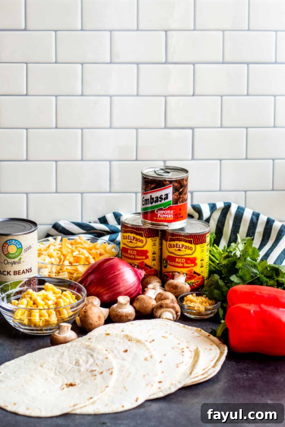 Assorted fresh ingredients for Mexican lasagna, including bell peppers, onions, mushrooms, tortillas, cheese, and beans, artfully arranged on a table.